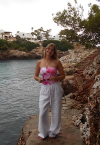 Jonge vrouw in een roze bloemen top en witte broek staat op een rotsachtige oever naast kalm water, met kusthuizen en groen in de achtergrond.