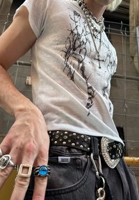 T-shirt branca com design gráfico preto, combinada com cinto preto com taxinhas, jeans de ganga e múltiplos anéis de prata. Colar de corrente prateada notável.