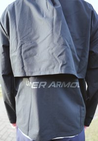 Black long-sleeve top with a layered design, made from a lightweight fabric, featuring a prominent silver "UNDER ARMOUR" logo on the back.