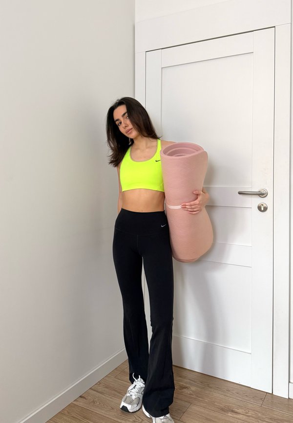 Woman in neon yellow sports bra and black leggings holding a rolled pink yoga mat standing indoors near a white door.