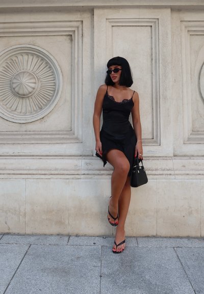 Vestido de satén negro con encaje, corsé ajustado y gran abertura. Chanclas negras, esmalte de uñas rojo y un pequeño bolso negro. Pared decorativa de fondo.