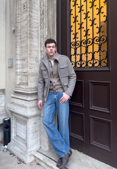 Joven con chaqueta beige de capas y vaqueros azules apoyado en una pared de piedra junto a una puerta de madera oscura con un panel de hierro ornamental.