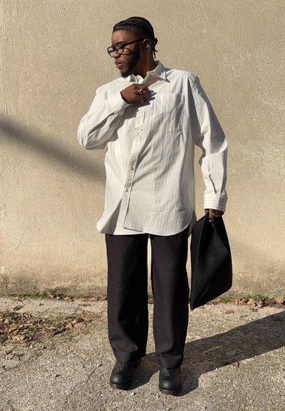 Camisa de manga larga blanca con rayas, pantalones de pierna ancha negros, zapatos negros y un bolso negro texturizado. Fondo neutro con sombras.