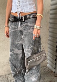 Šedé maskáčové široké džíny s černým opaskem se cvočky. Šedá kabelka se stříbrnými akcenty je držena v levé ruce spolu s různými náramky.