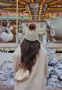 Gorro de malha bege com um pom-pom castanho, cabelo escuro longo, casaco creme e uma pequena bolsa bege. O fundo apresenta uma roda-gigante e árvores cobertas de neve.