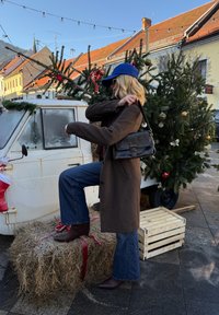 Manteau en laine marron, casquette bleue, sac à main noir, jean évasé, bottes bordeaux, posé sur une meule de foin avec des arbres décorés et un véhicule vintage.