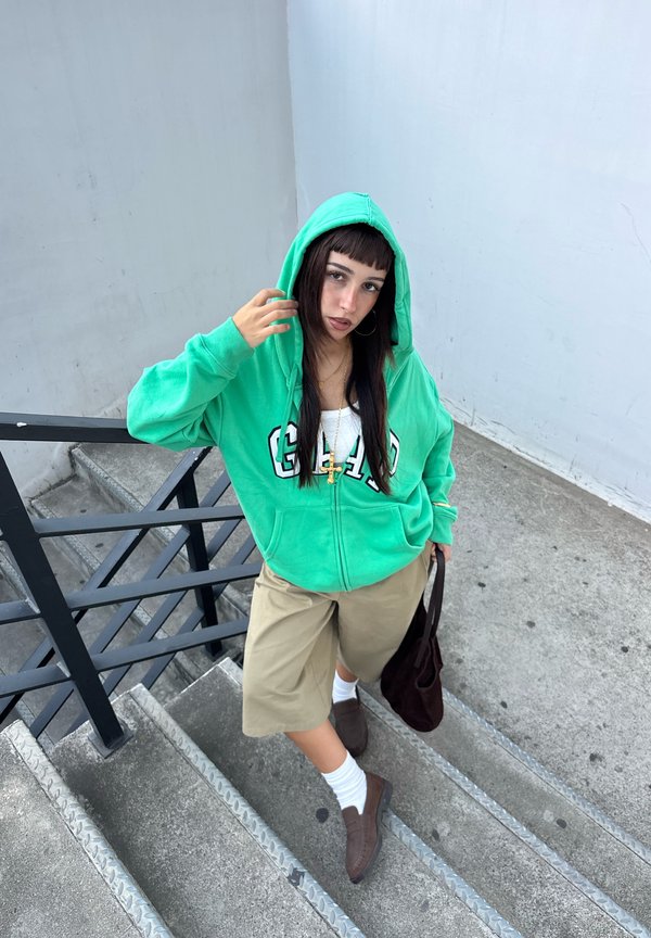 Une femme portant un sweat à capuche vert vif, un short beige, des chaussettes blanches et des chaussures brunes se tient sur des escaliers extérieurs et porte un sac fourre-tout sombre.