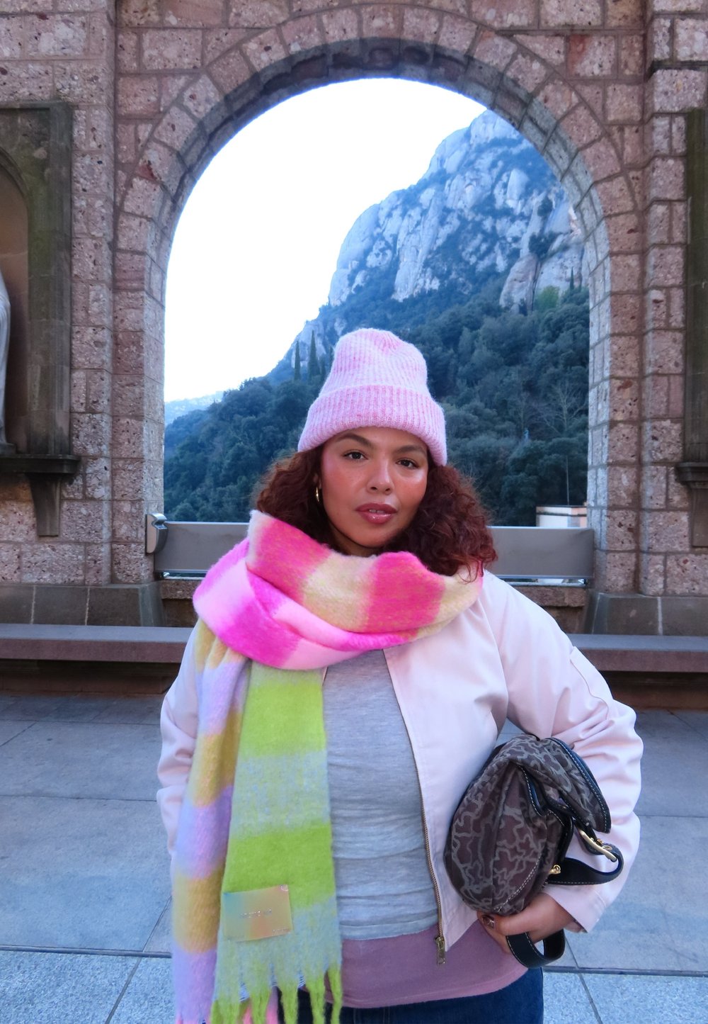 Femme portant un bonnet rose et une écharpe colorée, debout sous une arche en pierre avec des montagnes et une forêt en arrière-plan, tenant un sac à motifs.
