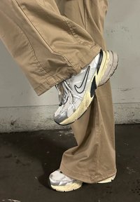 Ασημένια και λευκά αθλητικά παπούτσια με διχτυωτό επάνω μέρος, μαύρες λεπτομέρειες και χοντρές, μαλακές σόλες. Συνδυασμένα με φαρδιά καφέ παντελόνια.