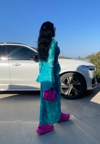 Transparent teal dress with frilled sleeves, paired with bright pink Crocs and a small purple bag. A white SUV is in the background.