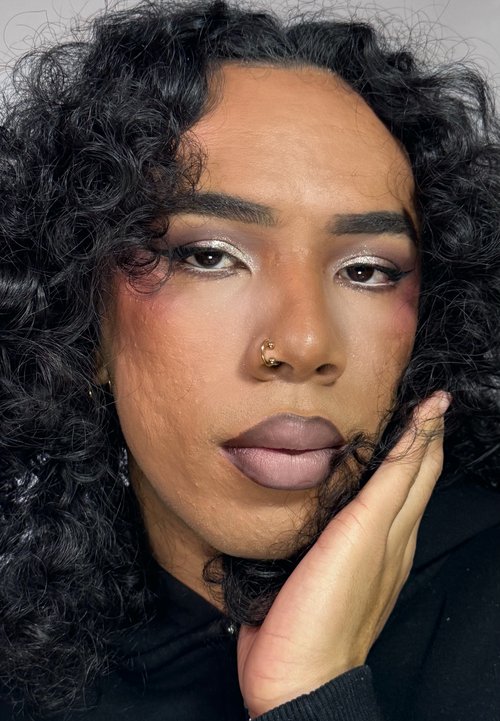 Primer plano de un rostro con cabello oscuro y rizado; maquillaje audaz con sombras de ojos verdes y púrpuras, labios marrones brillantes y un aro dorado en la nariz.