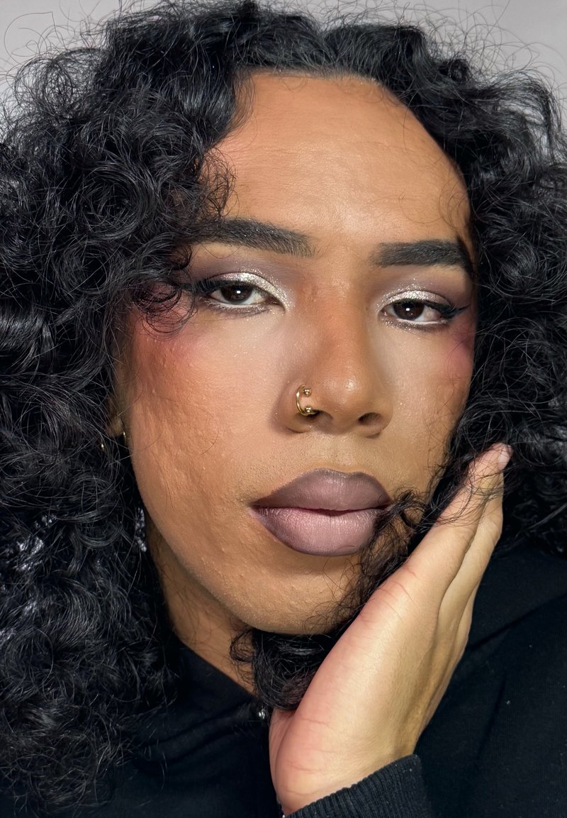 Primo piano di un volto con capelli ricci neri, un anello d'oro al naso, labbra nude opache e trucco occhi scintillante con accenti rosa e argento.