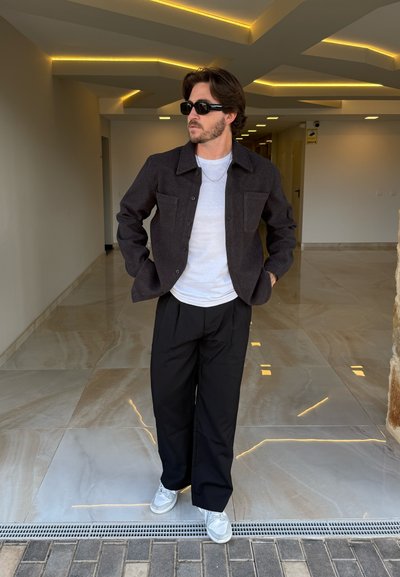 Chaqueta negra de botones sobre una camisa blanca, combinada con pantalones anchos negros y zapatillas grises. Suelo liso y pulido con acentos de luz.