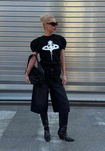 Camisa negra oversized con mangas abullonadas y diseño gráfico blanco, combinada con culottes de mezclilla negros anchos y botas de punta negra hasta la rodilla.