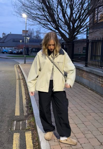 Chaqueta oversized crema con detalles negros, pantalones cargo negros y zuecos beiges. De pie sobre un camino de piedras en la suave luz de la tarde.