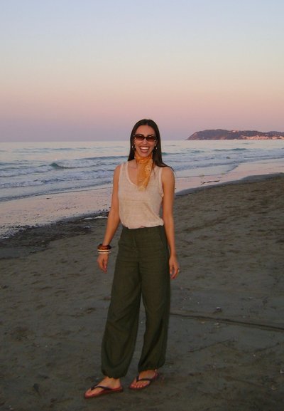 Top de tirantes color beige claro, pantalones de pierna ancha verde oliva, bufanda naranja, pulseras marrones y chanclas, de pie en una playa de arena al atardecer.