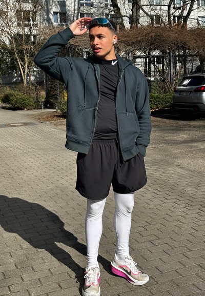 Atleta que lleva una sudadera con capucha de color azul oscuro, camiseta negra, pantalones cortos negros, mallas blancas y zapatillas grises con detalles en rosa, de pie al aire libre.