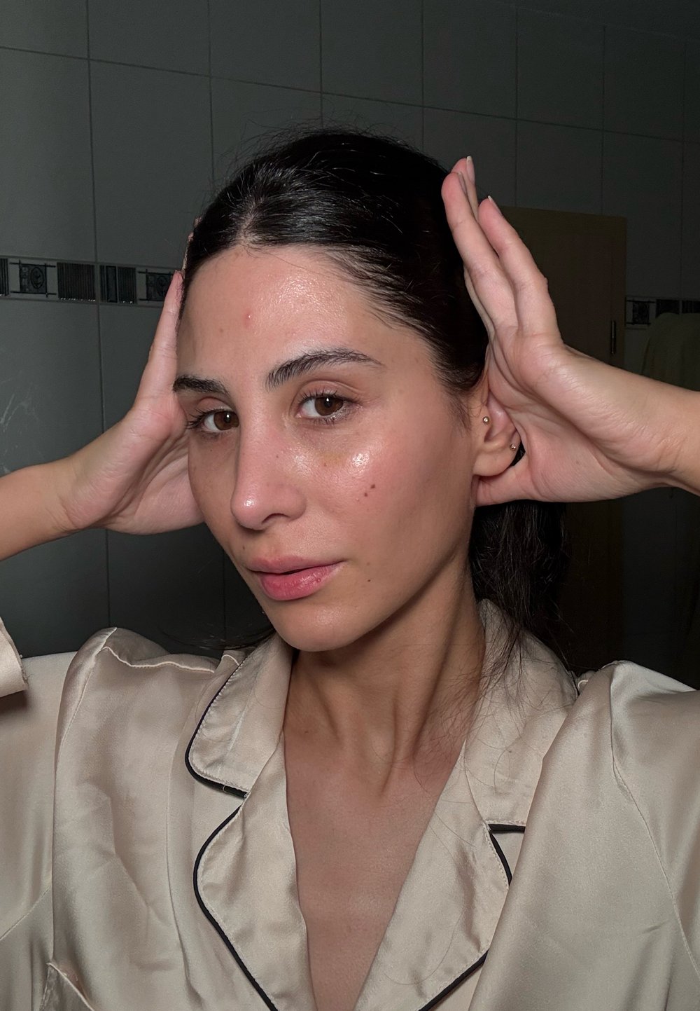Vrouw met lang, donker haar, gekleed in een lichtbeige satijnen top met zwarte bies, die haar hoofd met beide handen vasthoudt en een natuurlijke teint toont.