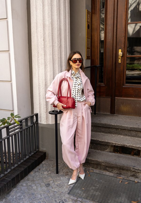Femeie îmbrăcată într-o ținută roz și tocuri albe stă lângă trepte de piatră, ținând o geantă roșie, purtând ochelari de soare și o bluză cu buline.