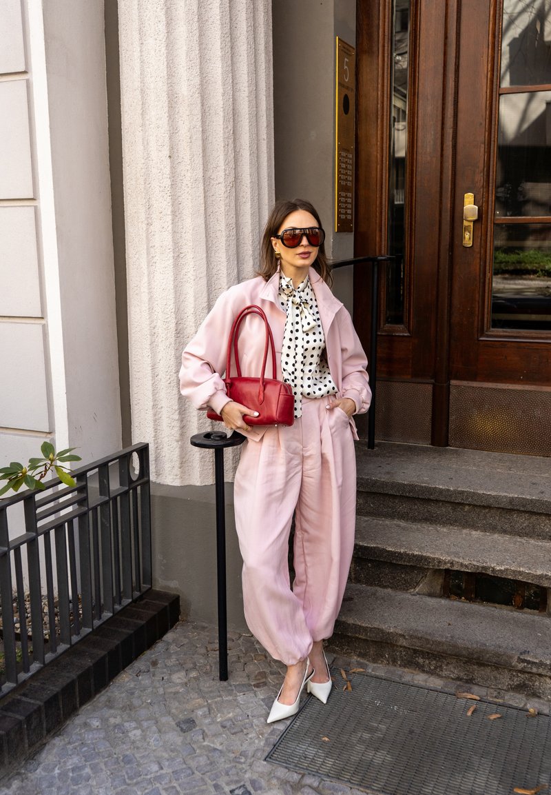 Femme en tenue rose et talons blancs debout près d'escaliers en pierre, tenant un sac à main rouge, portant des lunettes de soleil et un chemisier à pois.