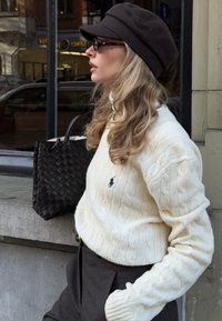 Γυναίκα που φοράει λευκό πουλόβερ με πλεκτό καλώδιο, σκούφο και παντελόνι σκούρου χρώματος, γυαλιά ηλίου και κρατάει μια μαύρη πλεκτή τσάντα δίπλα σε πέτρινο περβάζι.