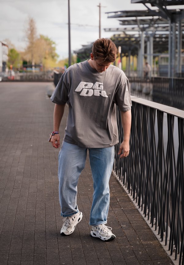 Camiseta gris de gran tamaño con logo blanco, jeans azul claro y zapatillas blancas. Persona caminando por un camino pavimentado al lado de una barandilla.