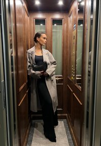 Casaco cinza oversized sobre uma blusa e calças pretas, segurando uma mala preta com tachas, de pé em um elevador de madeira com detalhes em metal.