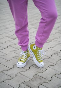 Keltaiset canvas-korkeavartiset tennarit, joissa on valkoiset kumipohjat ja nauhakiinnitys, yhdistettynä löysiin violetteihin verkkareihin. Taustalla on tekstuuripintainen asfaltti.