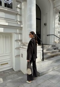 Zwarte oversized blazer en uitlopende broek, crèmekleurige stilettodollars, en een kleine zilveren handtas, staand op grijze stenen trappen tegen een witte architectuur.