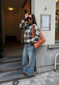 Maglione lavorato a maglia spessa in un motivo argyle teal e marrone, abbinato a jeans larghi sbiaditi. Porta una grande borsa a tracolla in pelle marrone.