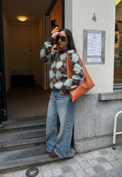 Suéter de punto grueso en color teal con un patrón argyle marrón, combinado con jeans de pierna ancha y desvanecidos. Lleva un gran bolso de cuero marrón.