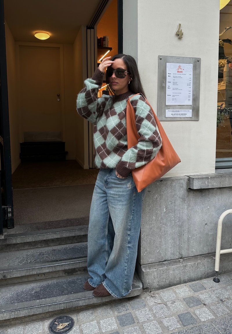 Pull en tricot épais dans un motif argyle teal et marron, associé à un jean large délavé. Elle porte un grand sac fourre-tout en cuir marron.