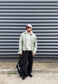 Svetlozelená bunda na zips cez sivú mikinu s kapucňou, čierne široké nohavice, čierna taška tote, sivá šiltovka a slnečné okuliare na pozadí textúrovanej sivej steny.