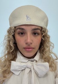 Berretto crema con logo canguro ricamato, abbinato a una camicetta bianca texturizzata con un grande colletto a fiocco. Riccioli naturali incorniciano il viso.