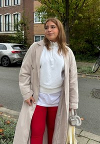 Ljus beige trenchcoat över en vit zip-up sweatshirt och röda leggings. Hon håller grå hörlurar och en beige tote-väska. Gatubakgrund.