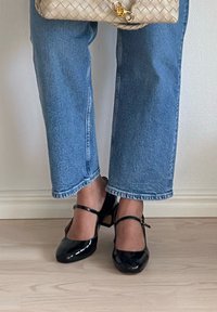 Schwarze Lackleder-Pumps mit Knöchelriemchen, quadratischer Zehenpartie. Kombiniert mit hellblauen, gerade geschnittenen Jeans, die den markanten Glanz der Schuhe zur Geltung bringen.