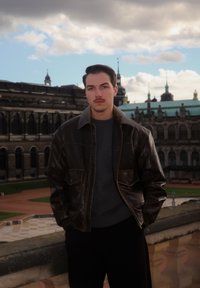 Casaco de couro castanho sobre um suéter cinzento, em pé contra um edifício histórico com grandes janelas e elementos decorativos. Céu nublado acima.