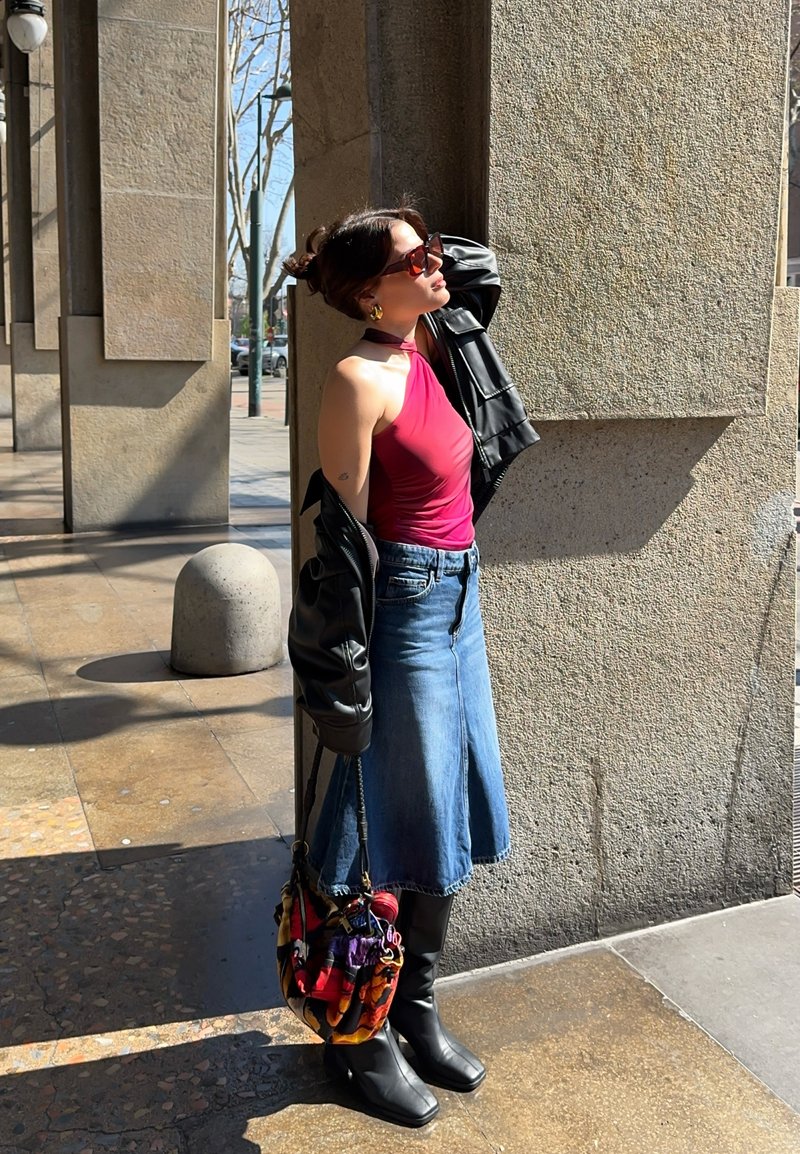 Frau mit Sonnenbrille, die ein rotes Neckholder-Top, Jeansrock, schwarze Stiefel und Lederjacke trägt und im Sonnenlicht an einer Steinsäule lehnt.