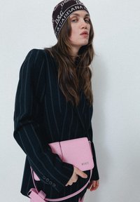 Blazer preto riscado, segurando uma pequena mala rosa a tiracolo. A mala apresenta uma textura suave e um detalhe em logótipo prateado. Gorro castanho com padrão.