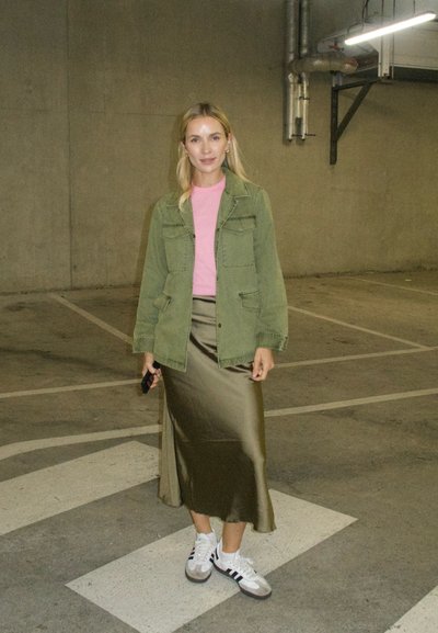 Chaqueta verde oliva, camiseta rosa, falda de satén verde oliva y zapatillas blancas con rayas negras. Fondo de hormigón con líneas de estacionamiento.