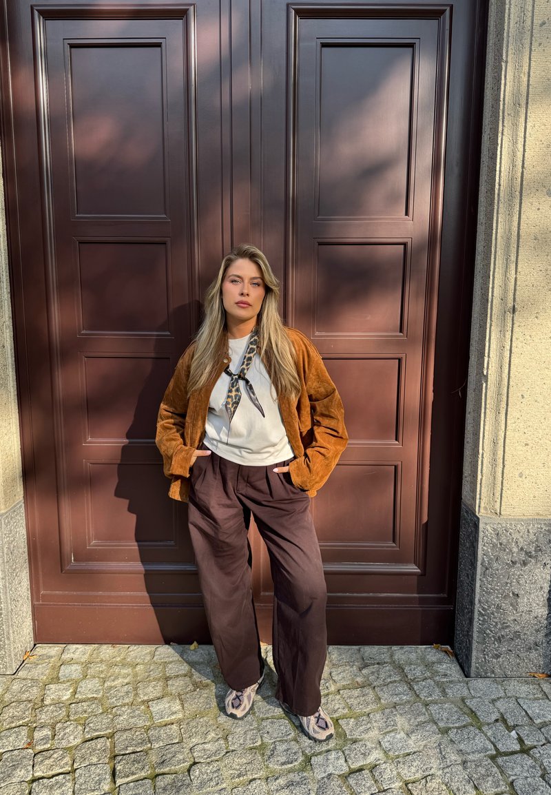 Brown suede jacket, white shirt, patterned scarf, loose brown trousers, and patterned shoes set against a dark wooden door and cobblestone background.
