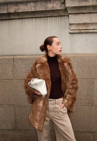 Bruine faux fur jas met een brede kraag, gedragen over een strakke bruine coltrui en crèmekleurige wijde broek, met een witte clutch tas in de hand.