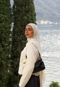 Hiyab blanco, chaqueta crema de doble botonadura con tejido texturizado, bolso negro con tachuelas, de pie junto al agua con árboles verdes de fondo.