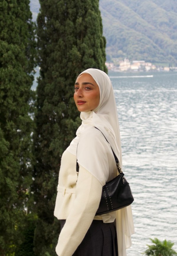 Witte hijab, crèmekleurige dubbelgeknoopte jas van textuurstof, zwarte handtas met studs, staand bij water met groene bomen op de achtergrond.