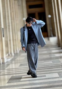 Hele sinine ülemisel bleiser, must T-särk, laiad hallid teksad, mustad jalanõud ja beret; aksessueeritud pruuni prillidega, jalutab koridoris.