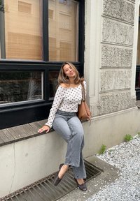 Biała bluzka z długim rękawem w brązowe kropki, wysokie szare dżinsy, brązowa torebka i brązowe buty. Siedzi na drewnianej półce.