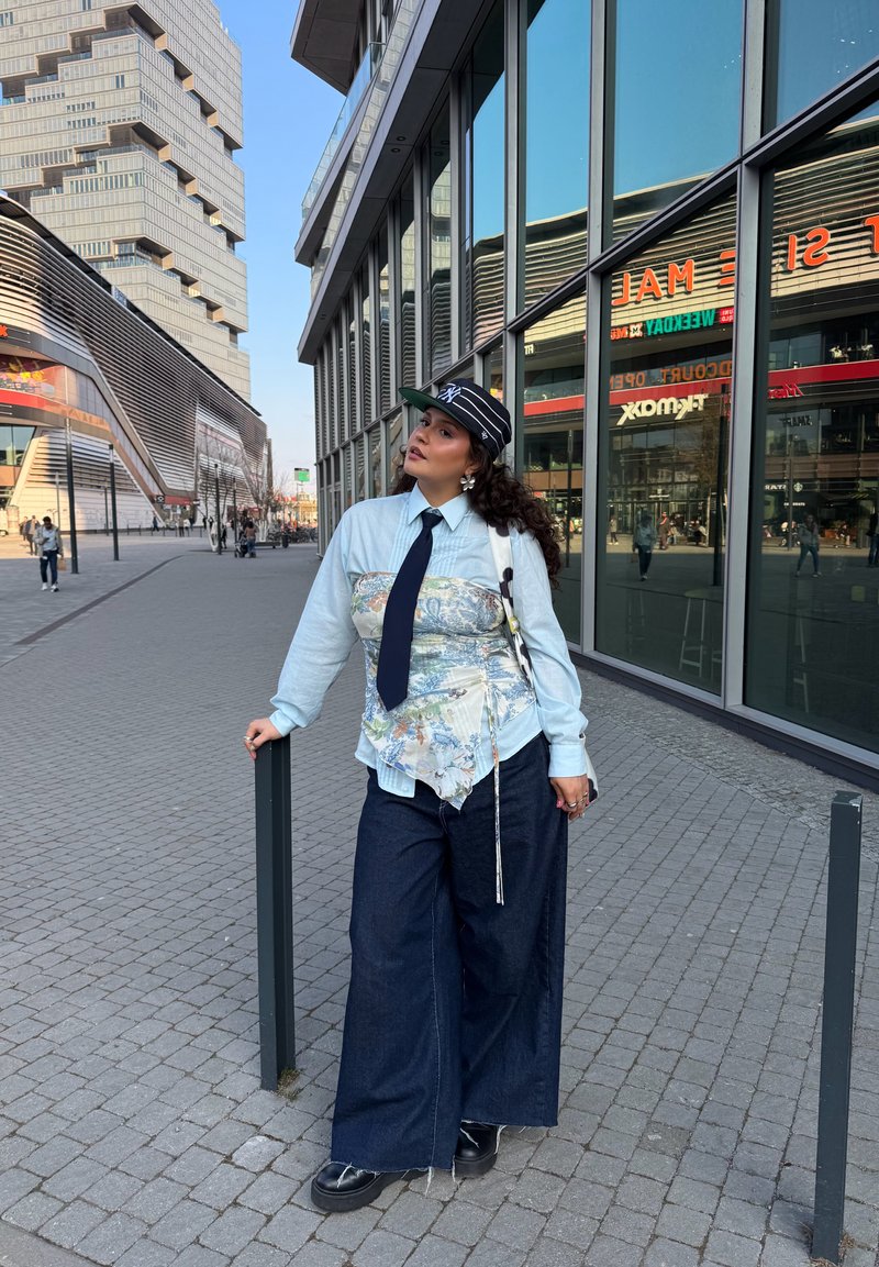 Žena odjevena u traperice širokih nogavica, cvjetni korzet preko plave košulje, mornarsko plava kravata i prugasta kapa, naslonjena na stup na gradskom pločniku.