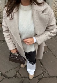 Femme portant un manteau beige, un pull blanc duveteux, un pantalon blanc, des chaussures beiges, tenant un sac à main marron foncé et montrant des bagues à sa main.