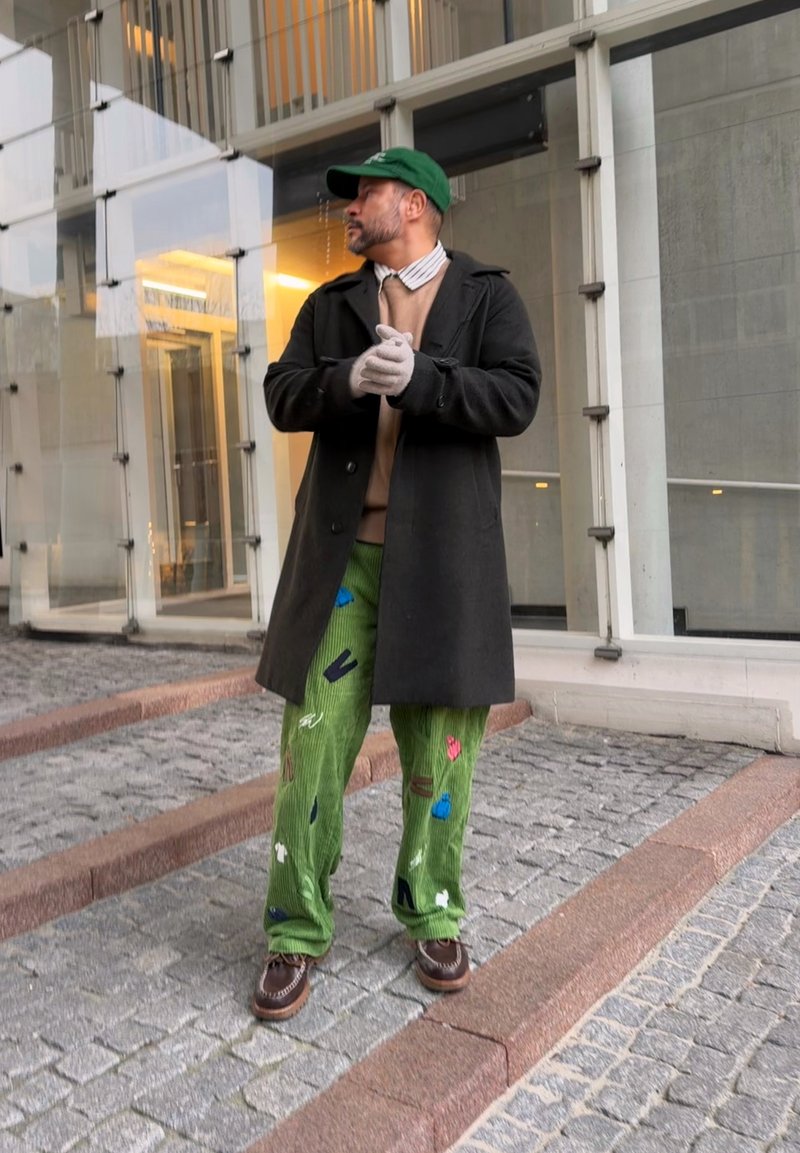 Homme portant une casquette verte, un manteau noir, un pull beige, un pantalon vert à motifs et des gants, debout sur un pavé en pierre à côté d'un bâtiment en verre.