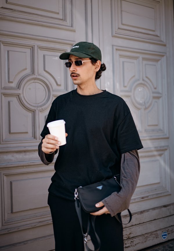 Schwarzes oversized T-Shirt, grauer langärmliger Unterzieher, schwarze Umhängetasche, grüne Kappe, Sonnenbrille, hält eine weiße Kaffeetasse vor einer Tür.